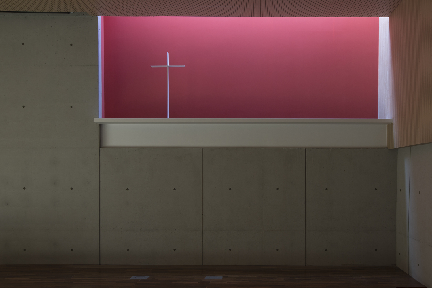 Interior chapel wall with high clerestory window revealing a pink-lit cross against the sky