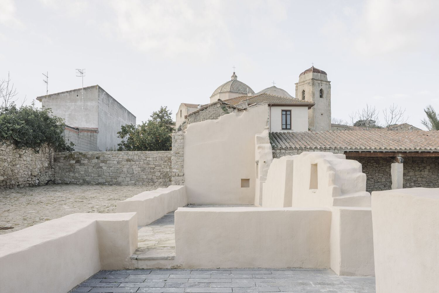 Cream stucco platforms and steps in a courtyard with a domed church and stone walls beyond