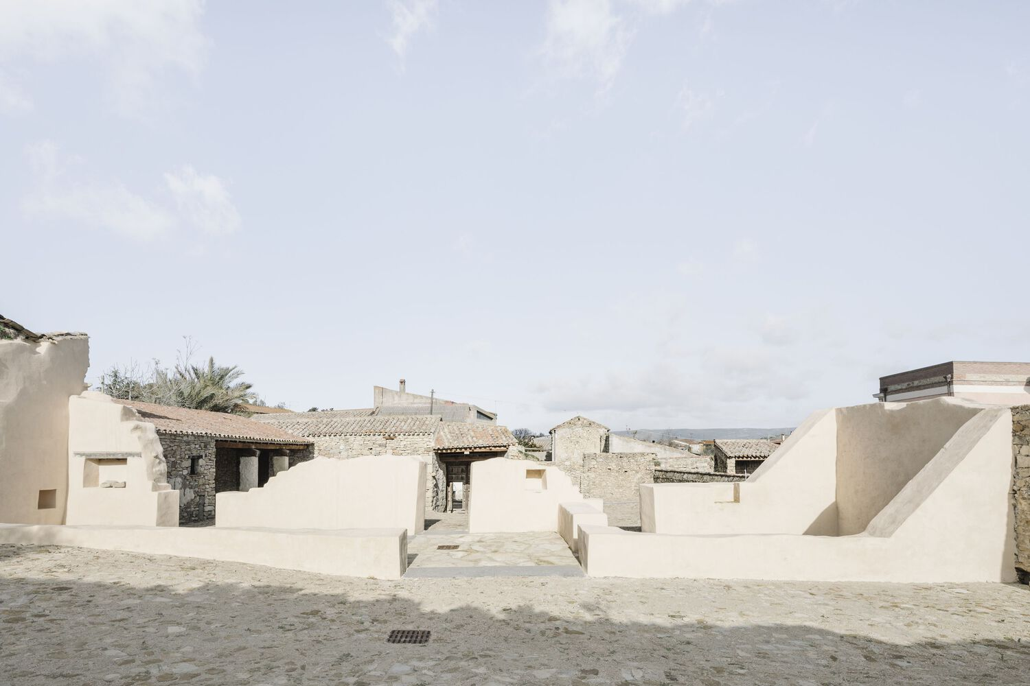Wide courtyard view with stepped stucco seating platforms facing stone ruins under an overcast sky