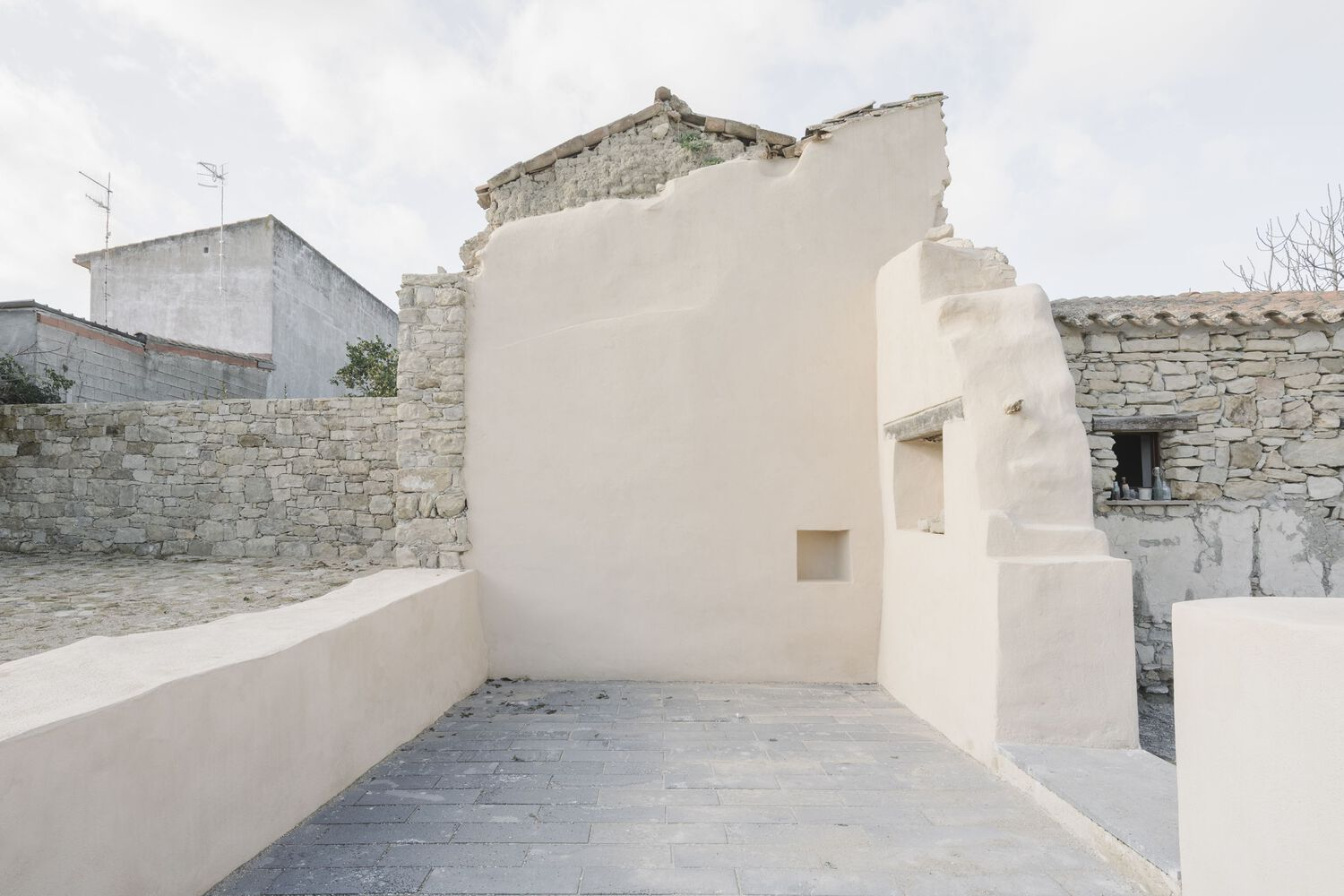 Cream stucco wall with small square openings beside a crumbling stone structure under a cloudy sky