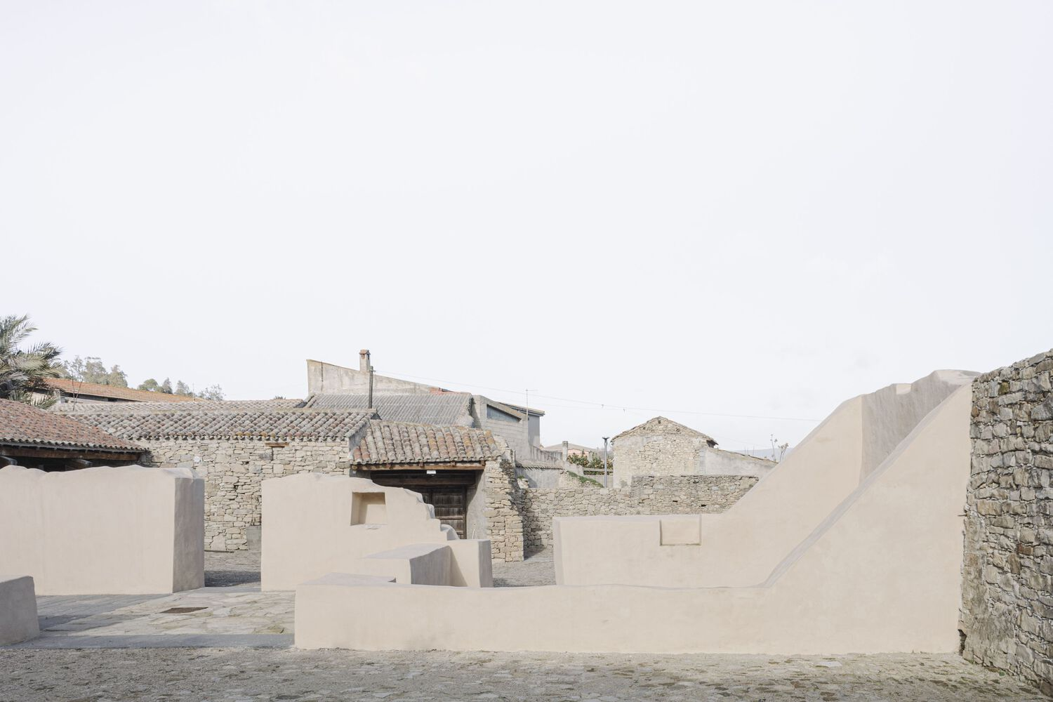Sculptural plastered walls forming stepped platforms in a courtyard framed by stone buildings with tile roofs