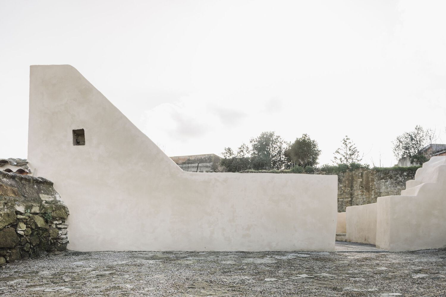 Sloped white rendered wall with small window opening framed by stone ruins and vegetation