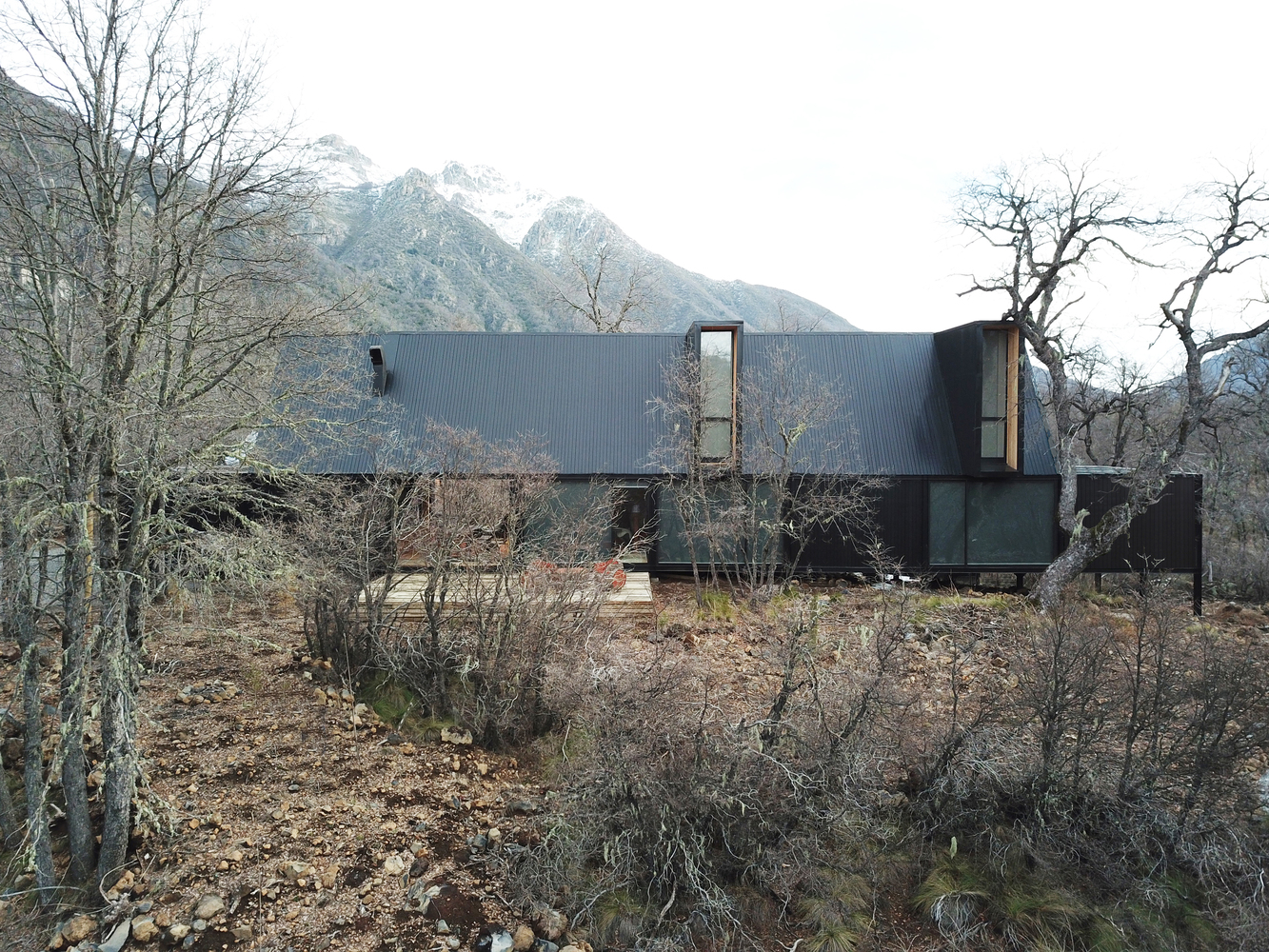Linear metal-clad volumes on a rocky hillside with bare winter trees and snow-capped mountains beyond
