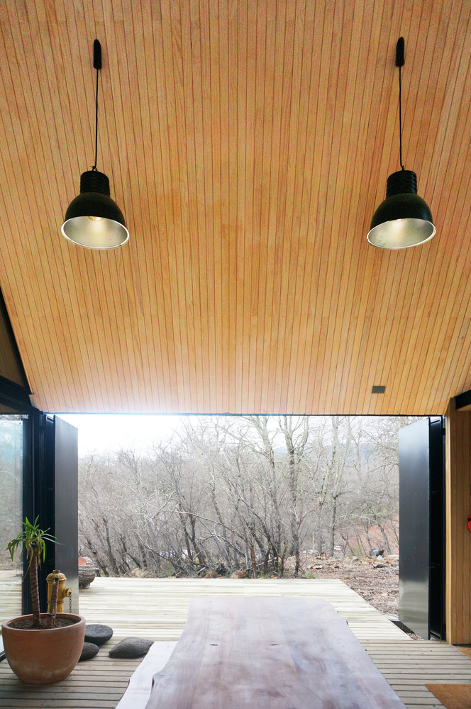View through the open corner glazing to the winter forest with two black pendant lights hanging from the wood ceiling