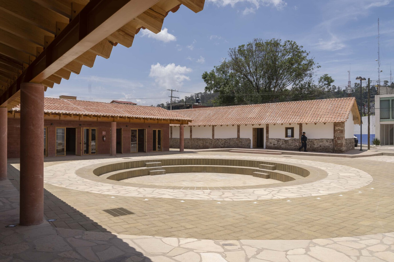 Central courtyard with circular stepped fountain basin surrounded by timber colonnade and tile-roofed buildings under blue sky
