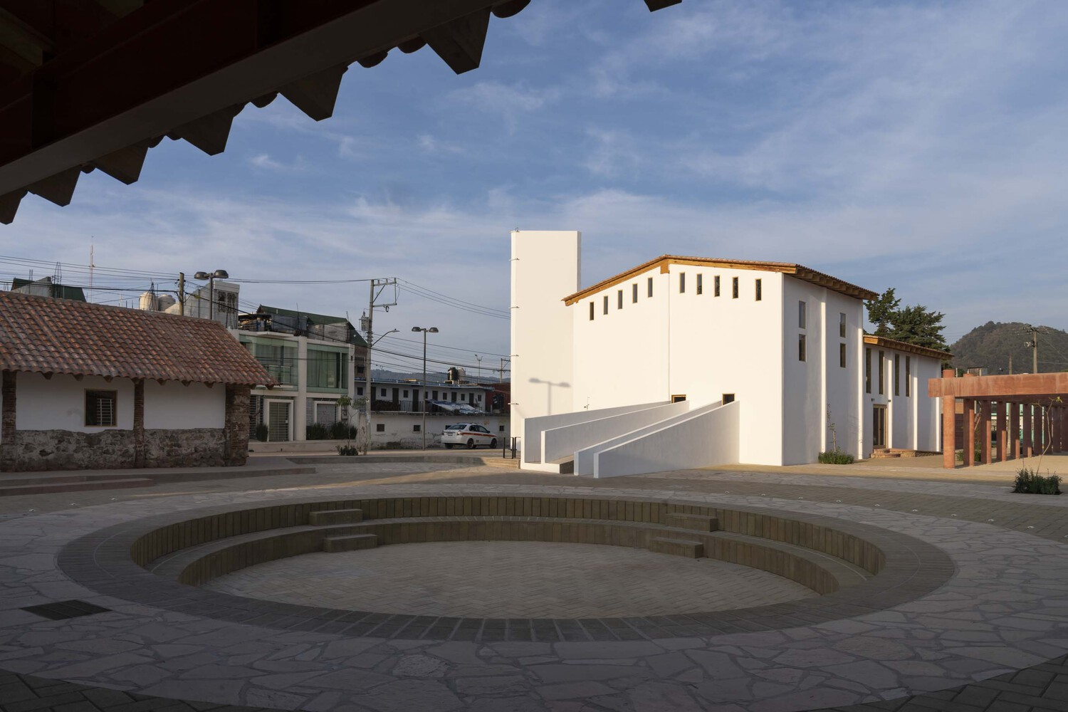Sunken circular plaza with stepped brick rings beside a white two-story pavilion under a partly cloudy sky