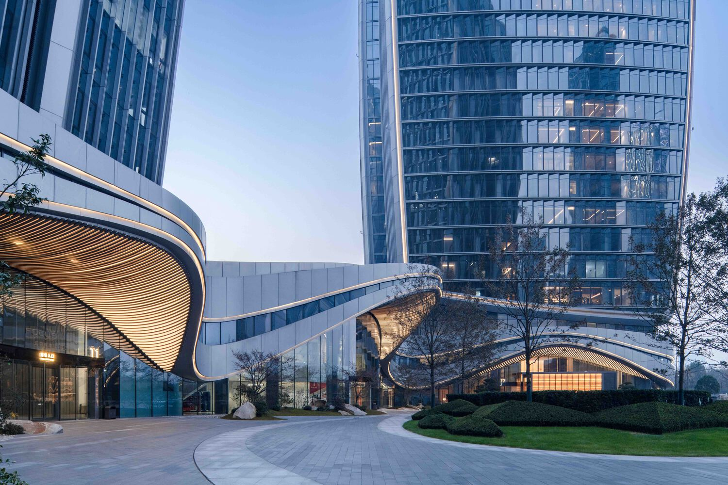 Curved glass tower base with wood-lined soffit and cantilevered canopy at dusk