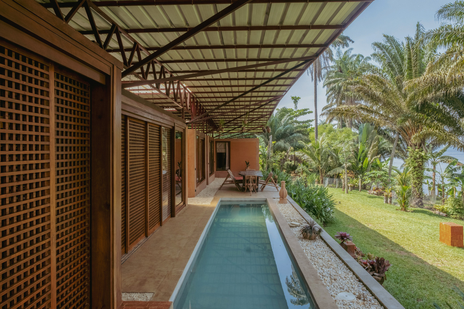 Lap pool alongside lattice-screened veranda with steel truss roof overlooking sloped lawn and palms