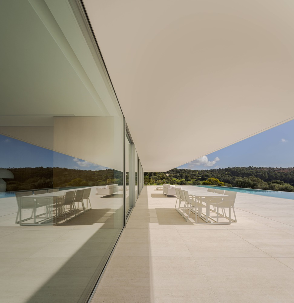 Outdoor terrace with white dining furniture beneath a deep overhang and reflected in the glazing