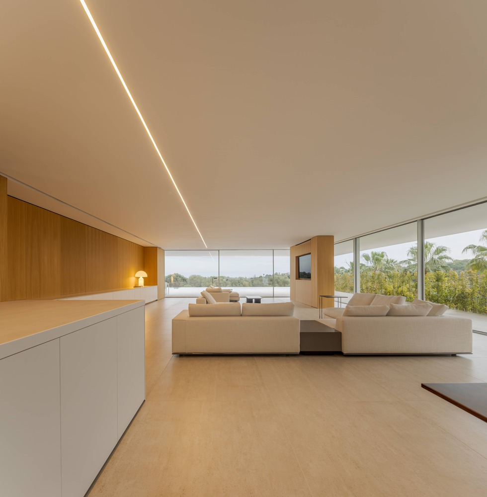 Open living room with continuous ceiling light slot and floor-to-ceiling glazing overlooking tropical landscape
