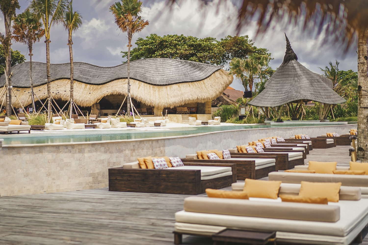 Pool deck with rows of loungers facing undulating thatch-roofed structures and palm trees