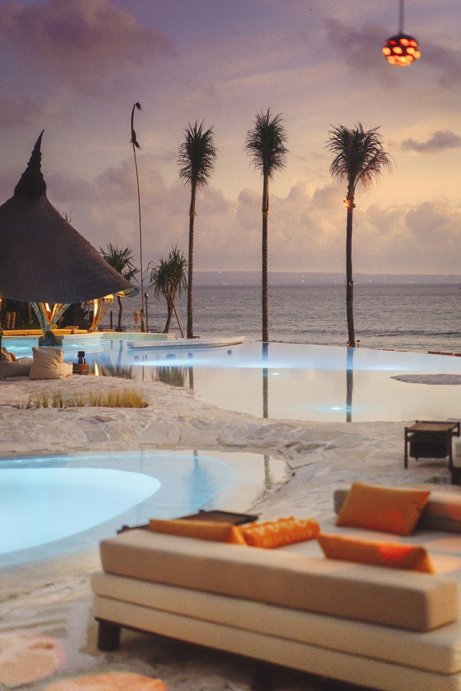 Infinity pool edge at dusk with thatched pavilion and palms silhouetted against the ocean