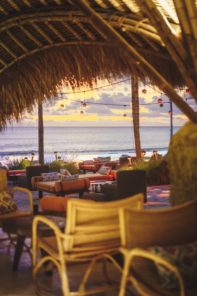 View through a thatch-roofed pavilion toward ocean and sunset sky with suspended pendant lights