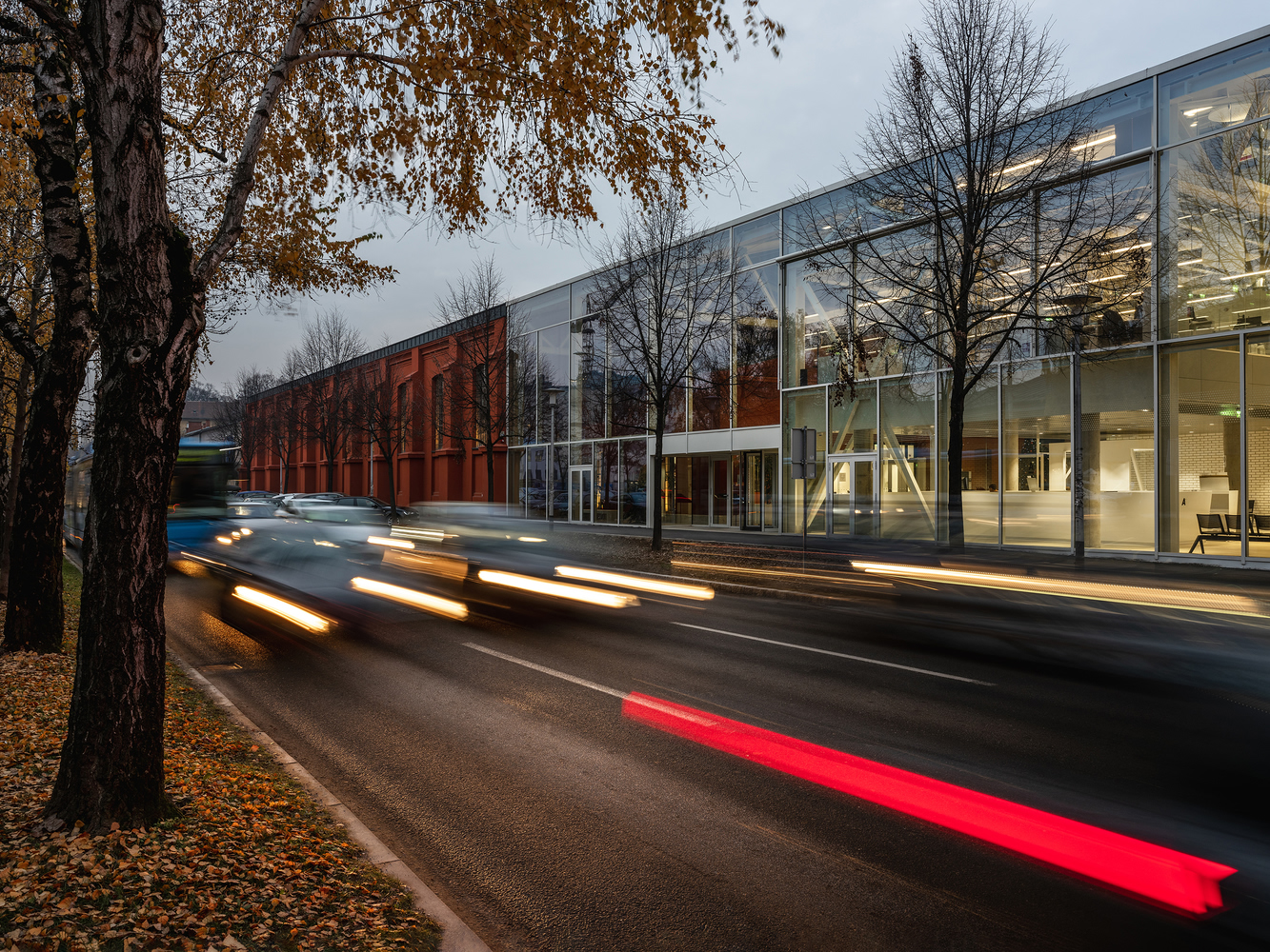 Streetside facade with glass curtain wall and red brick volume at dusk with vehicle light trails