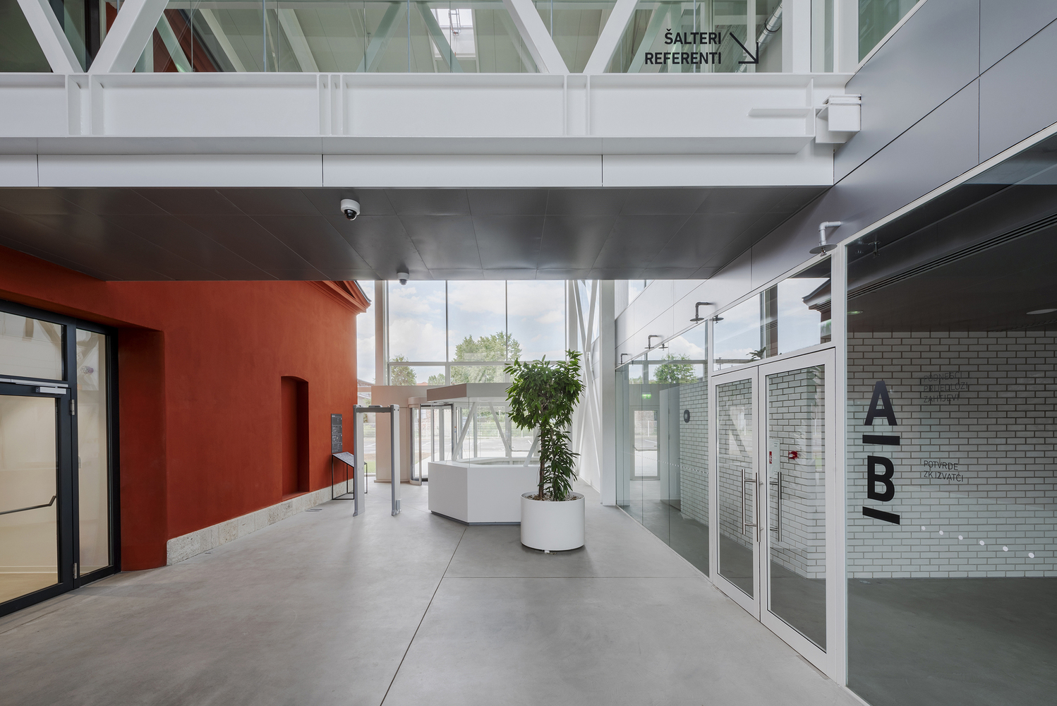 Interior corridor with orange wall panel, glazed partitions, and planted greenery under exposed ceiling structure