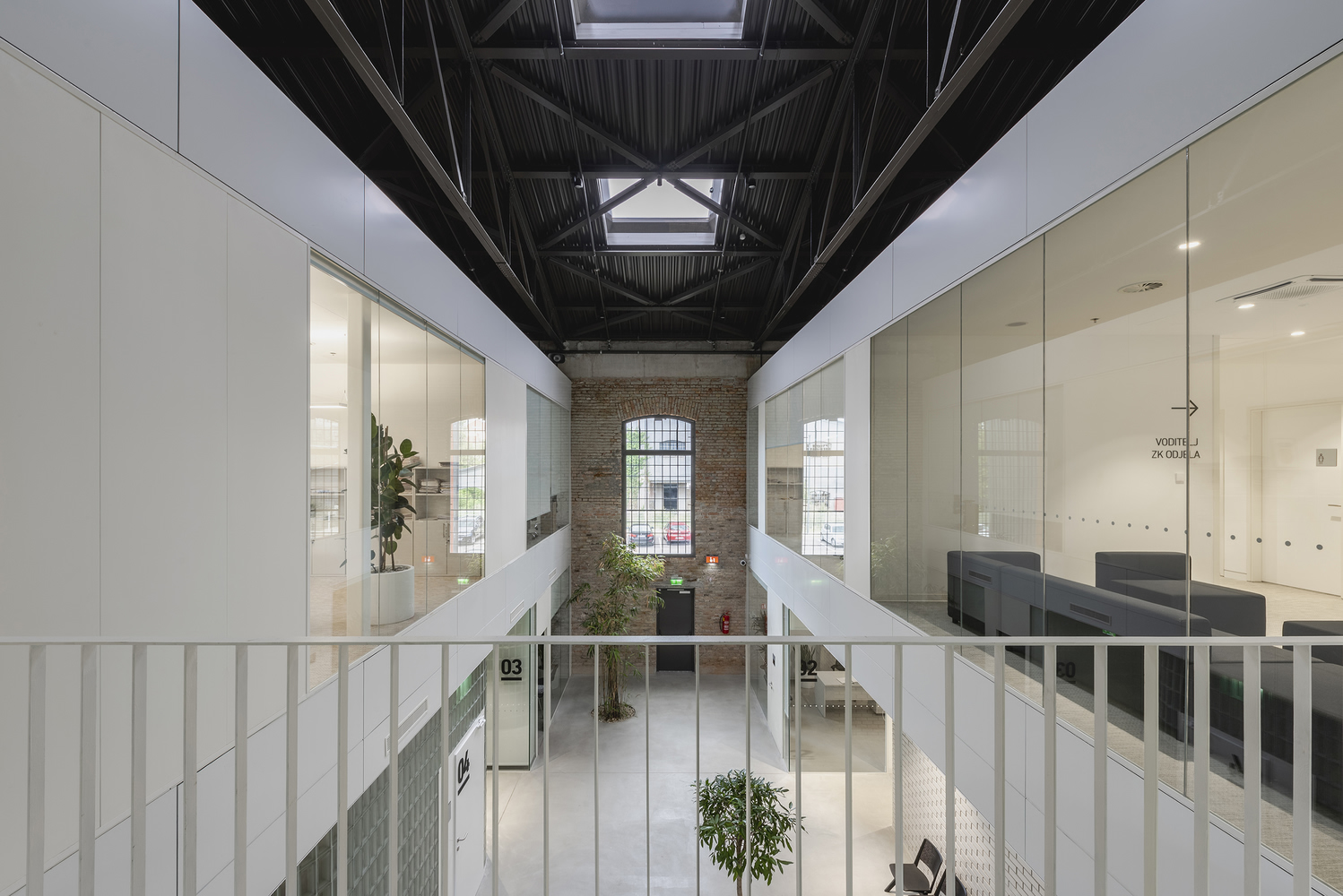 View across mezzanine bridge toward arched brick window and glazed office volumes with white panel balustrades