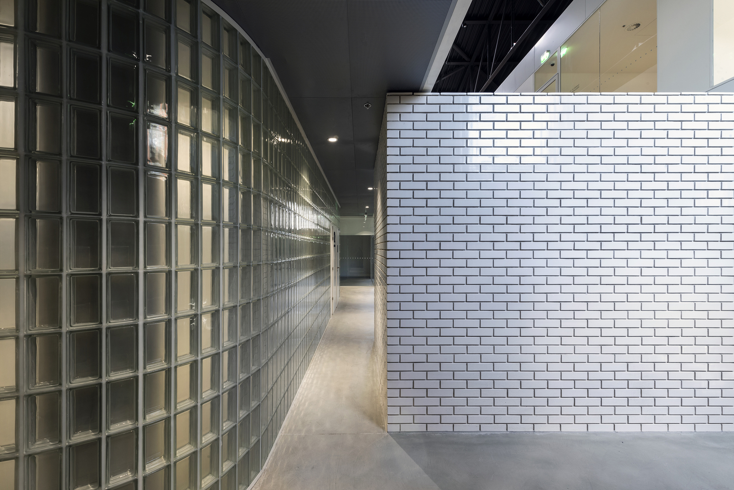 Interior corridor with curved glass block wall opposite white subway tile partition under black ceiling