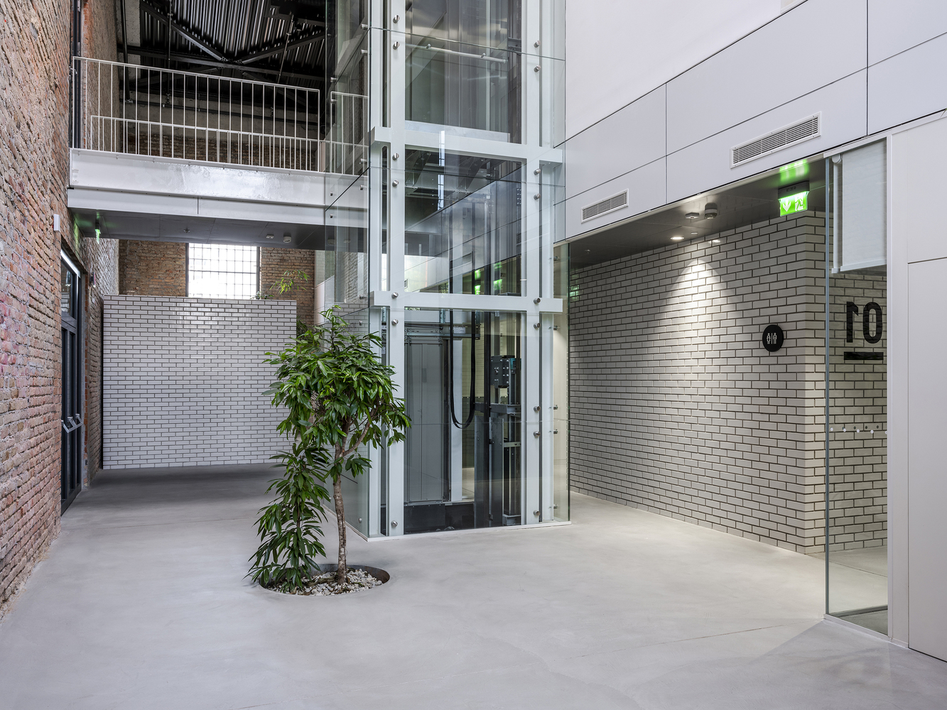 Glass elevator shaft and polished concrete floor with potted tree beside white tile entry wall