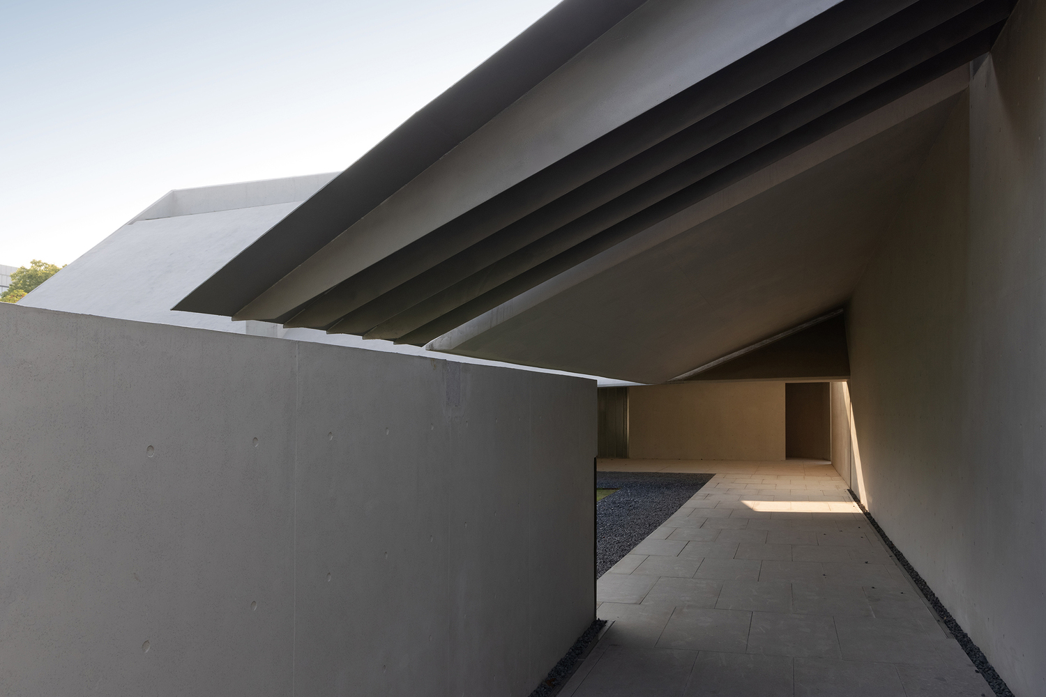 Covered walkway with ribbed concrete soffit casting shadows over stone paving at dusk