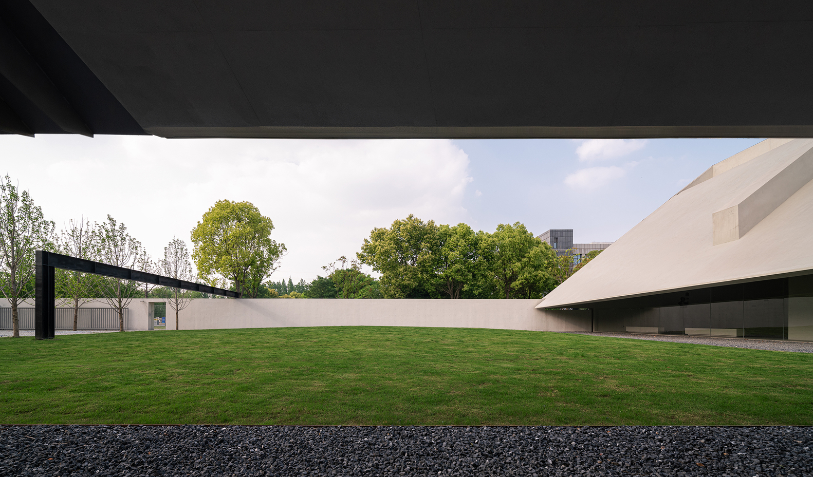 View across lawn toward long concrete wall with black canopy and angular white roof beyond