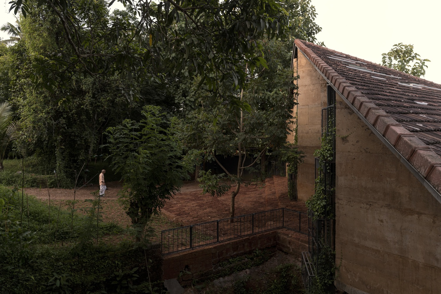 Gabled terracotta roof and rammed earth facade overlooking a courtyard with young trees and a person walking