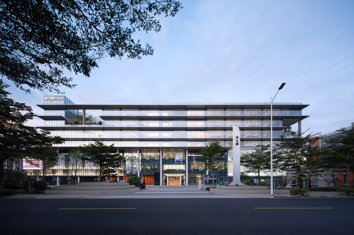 Street view of the horizontally banded glass and panel facade framed by roadside trees at dusk