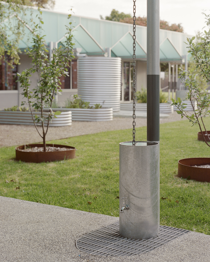 Concrete rainwater chain column with tap and steel grate in a lawn with planted beds and corrugated metal beyond