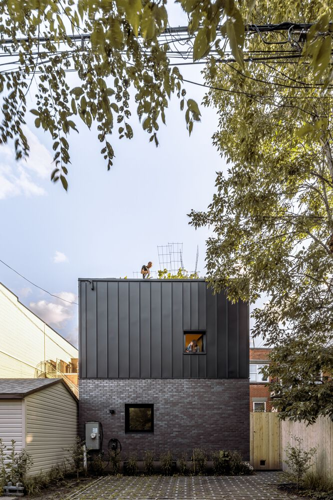 Street view of the two-story volume with dark metal cladding over brick base beneath overhanging birch branches