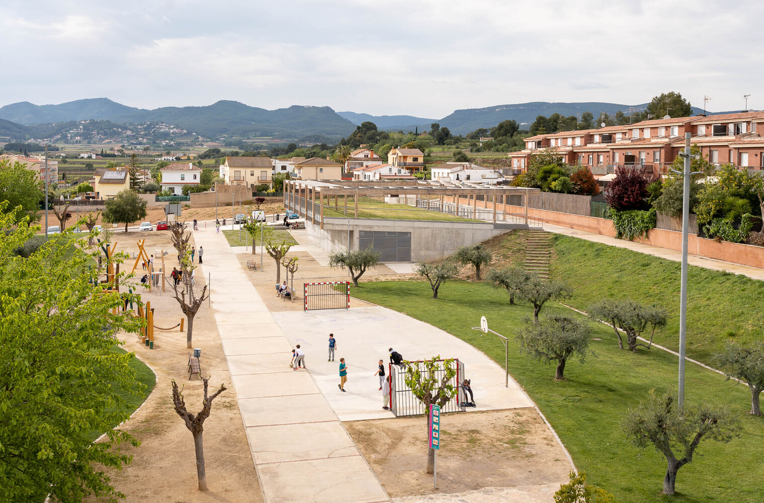 Elevated view of the public plaza with playgrounds, pruned trees and mountains visible in the distance