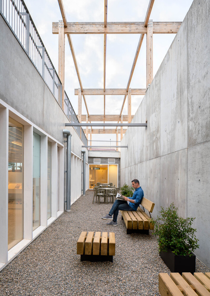 Gravel courtyard flanked by concrete walls with a timber pergola overhead and someone seated on a bench