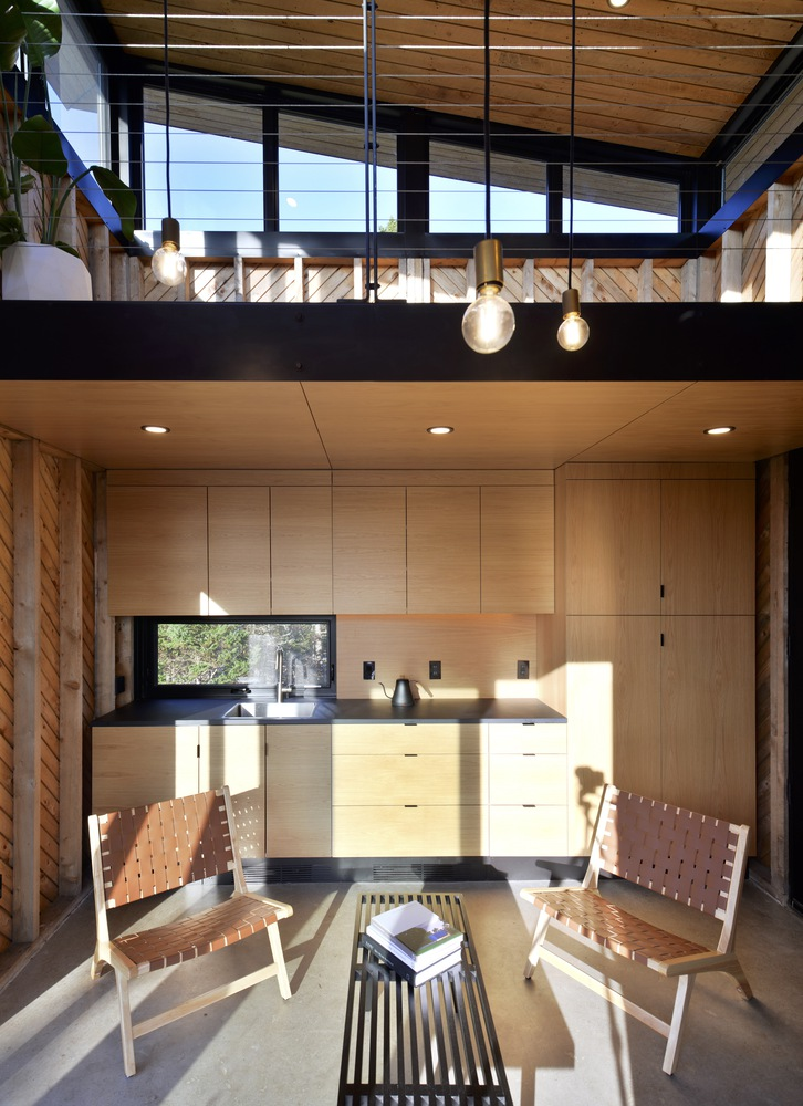 Double-height living space with plywood cabinetry, woven lounge chairs, and black steel mezzanine railing above
