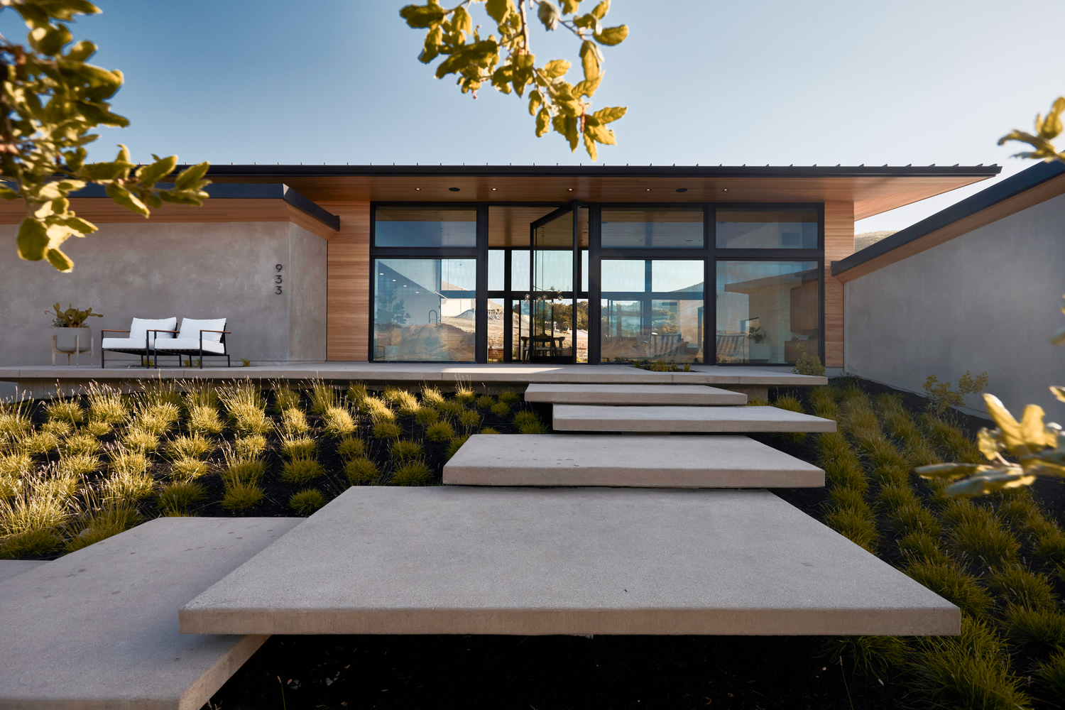 Floating concrete steps leading through planted grasses toward a glazed pavilion under afternoon light