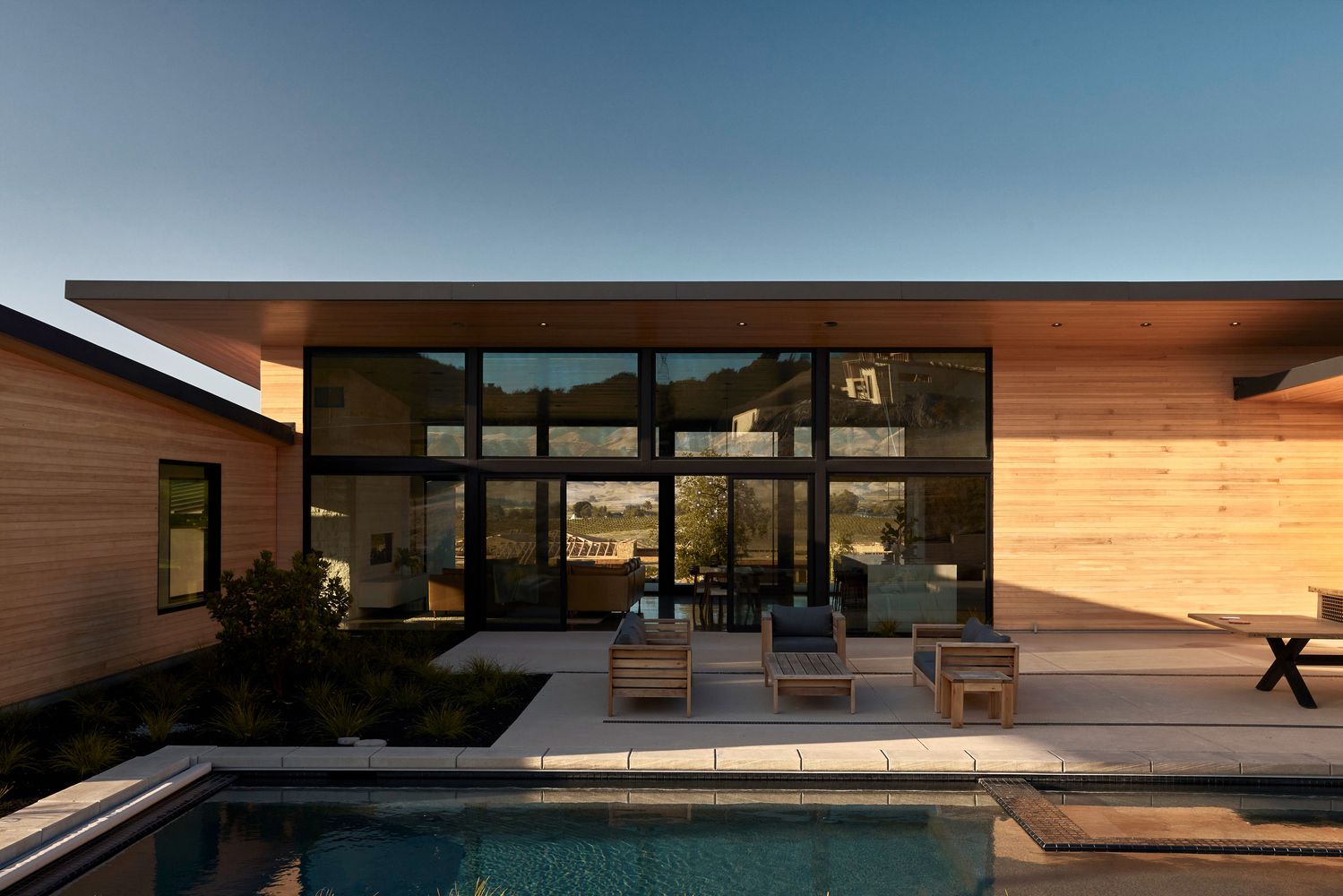 Rear courtyard with floor-to-ceiling glazing and timber lounge chairs beside the pool at twilight