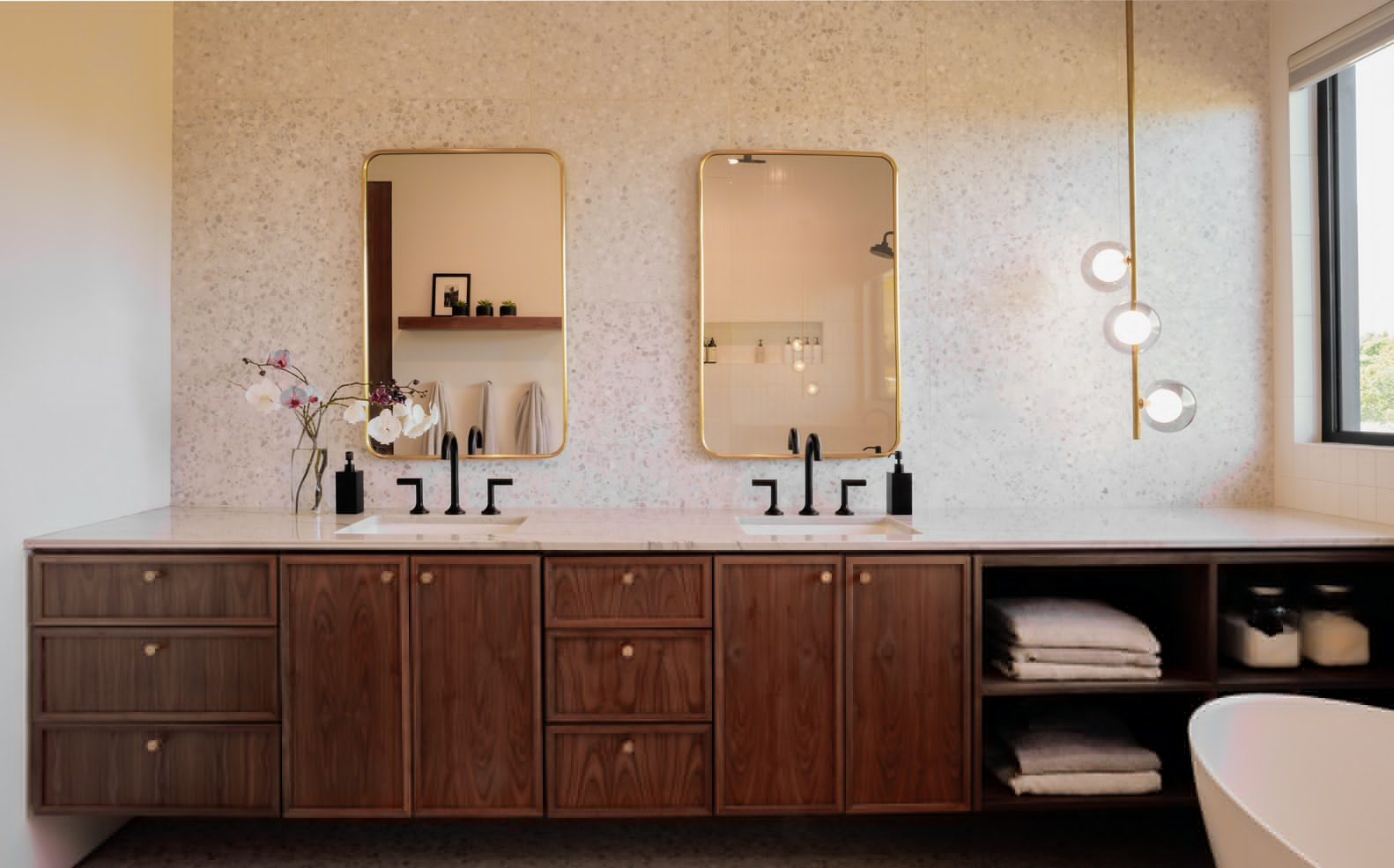 Double walnut vanity with brass-framed mirrors against a terrazzo wall in a bathroom