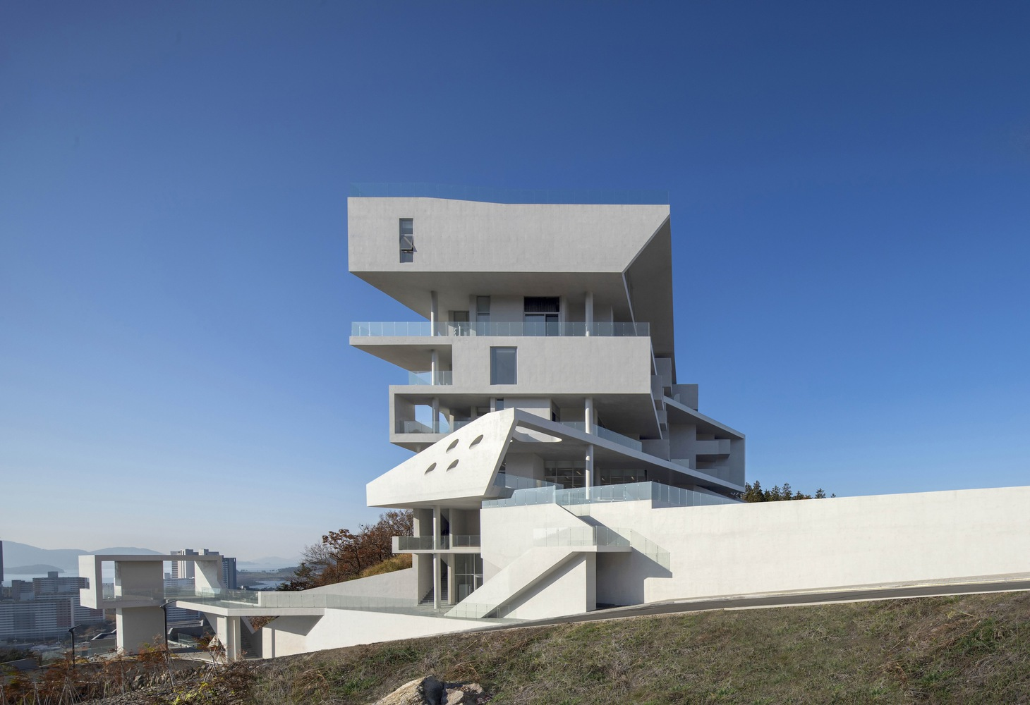 White concrete tower with offset floor plates and terraces rising above a hillside slope