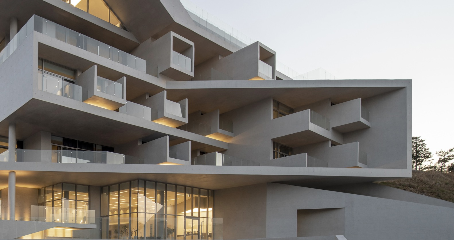 Stacked concrete slabs with recessed balconies illuminated from within at dusk