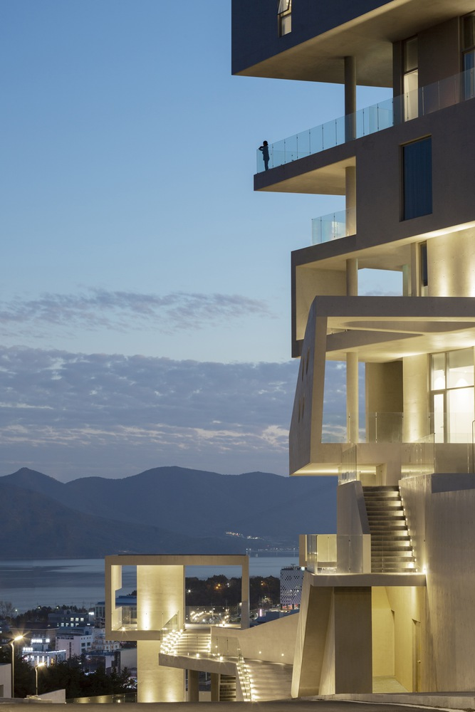 Illuminated stair volumes threading through stacked concrete slabs at twilight with a figure on upper terrace
