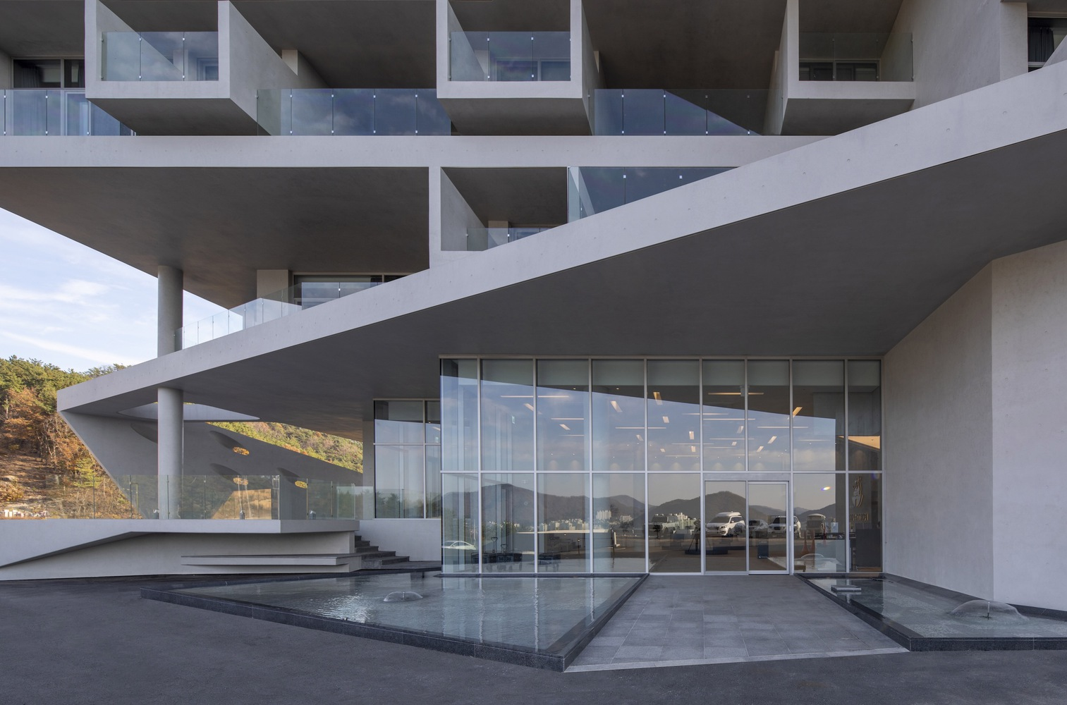 Ground level view of stacked concrete slabs with glass walls and a shallow reflecting pool at dusk