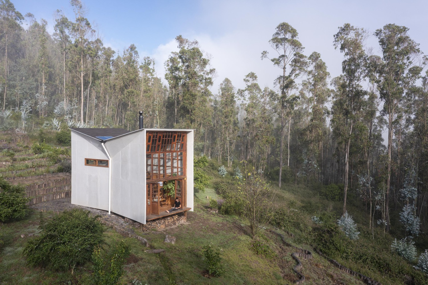 Elevated view of the white-clad cabin with timber-framed glass facade set among eucalyptus trees on a hillside