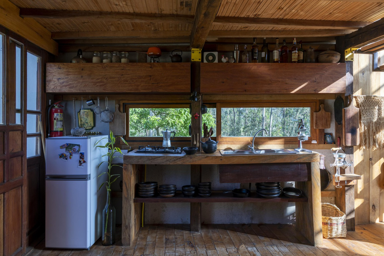 Kitchen area with exposed timber beams and open shelving beneath a horizontal window facing the forest