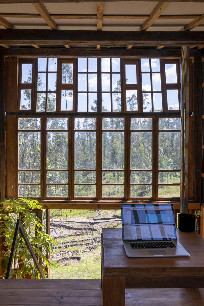 Desk with laptop beneath a timber-framed gridded window wall looking out to the sloped landscape and trees