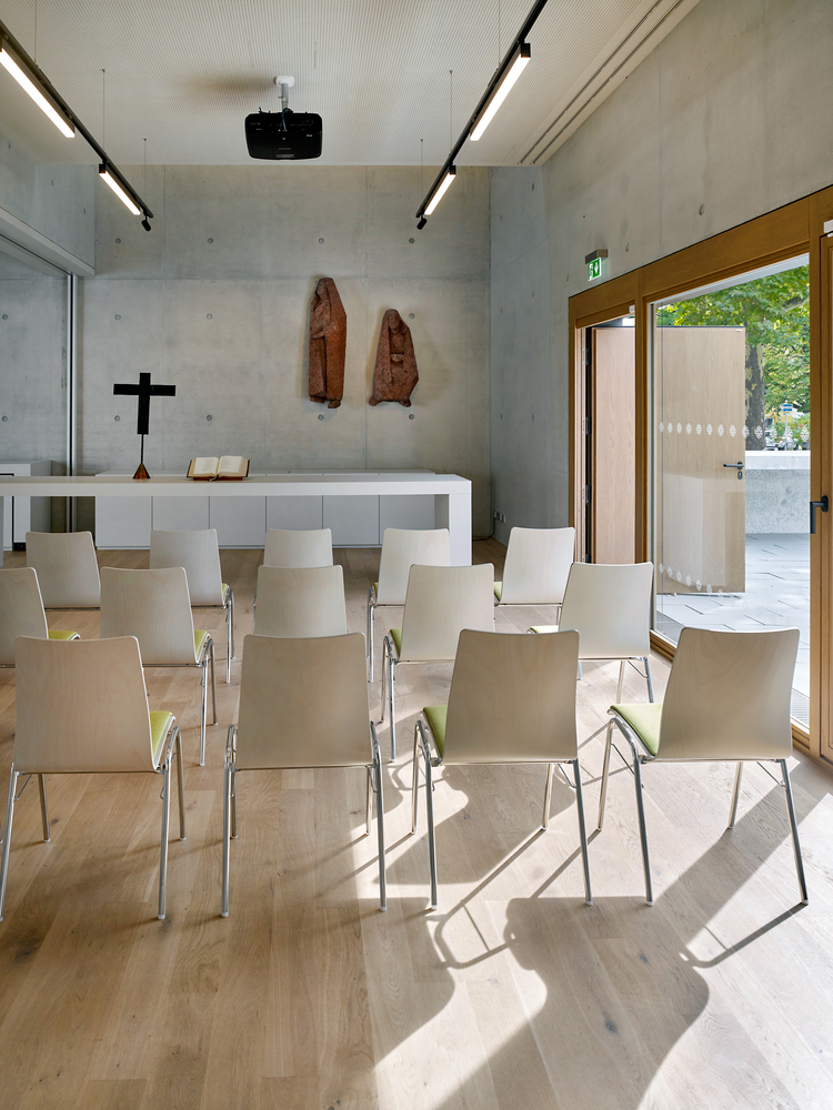 Interior chapel space with board-formed concrete walls, rows of light wood seating, and sunlight falling across the floor