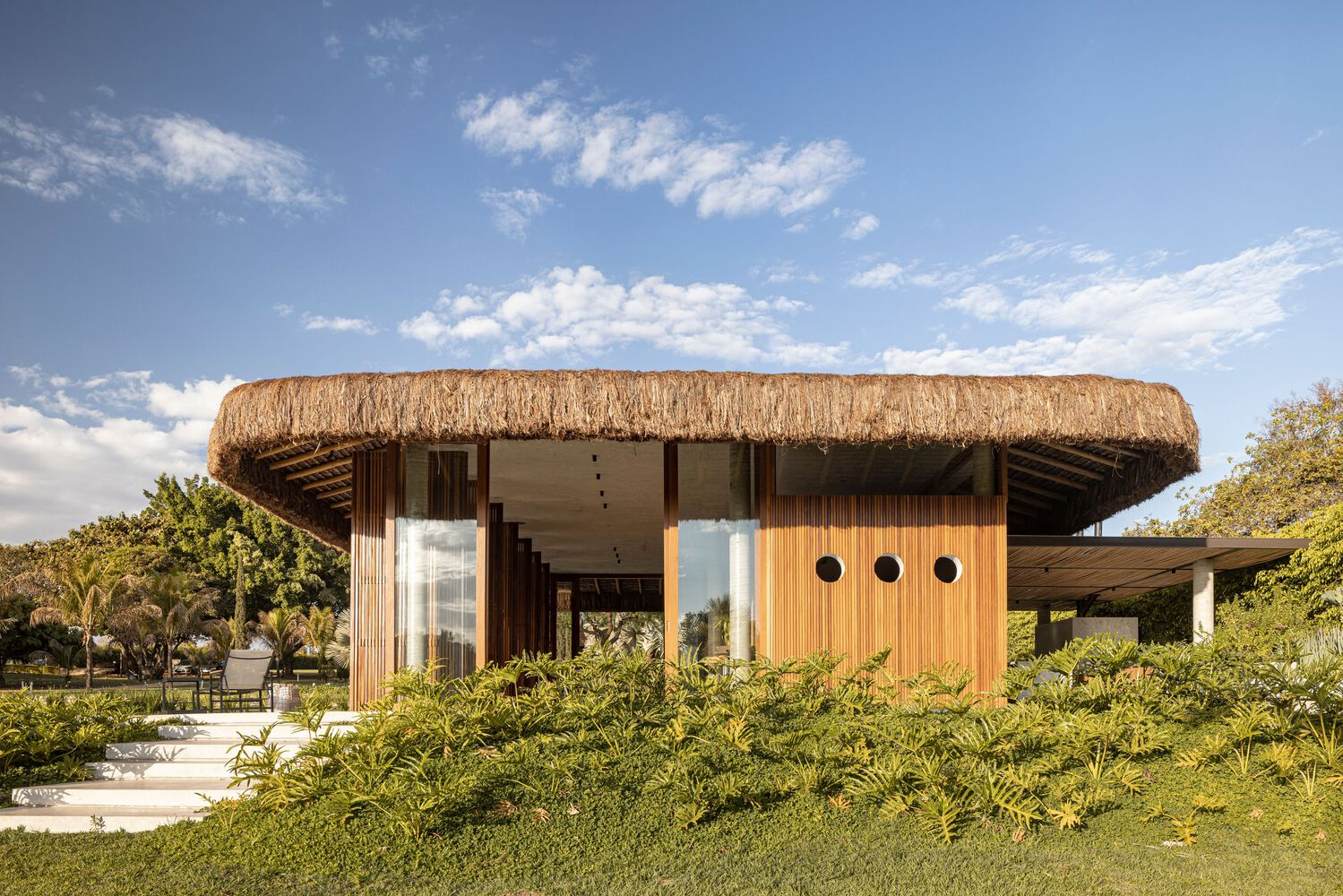 Thatched roof pavilion with timber and vertical wood cladding set within a planted lawn under afternoon clouds