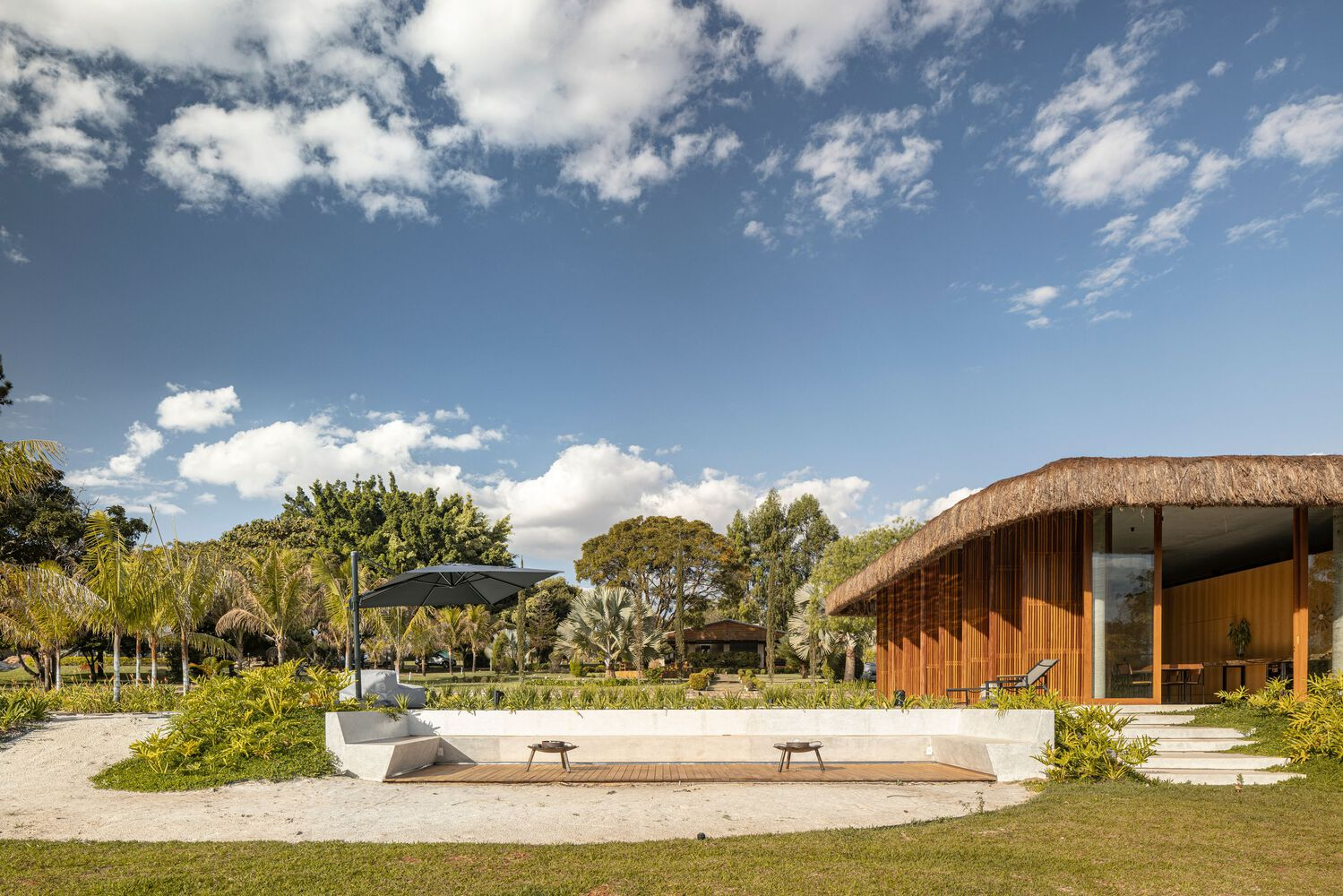 Timber pavilion with thatched roof beside a paved terrace and lawn under scattered clouds