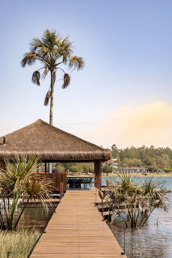 Timber boardwalk leading to a thatched roof gazebo at the water's edge with a palm tree overhead