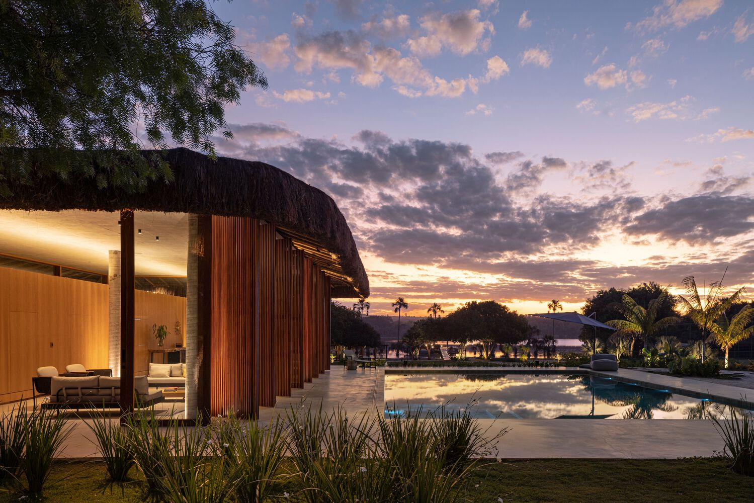 Curved thatched roof pavilion beside a reflecting pool at dusk with palm trees silhouetted against the sky