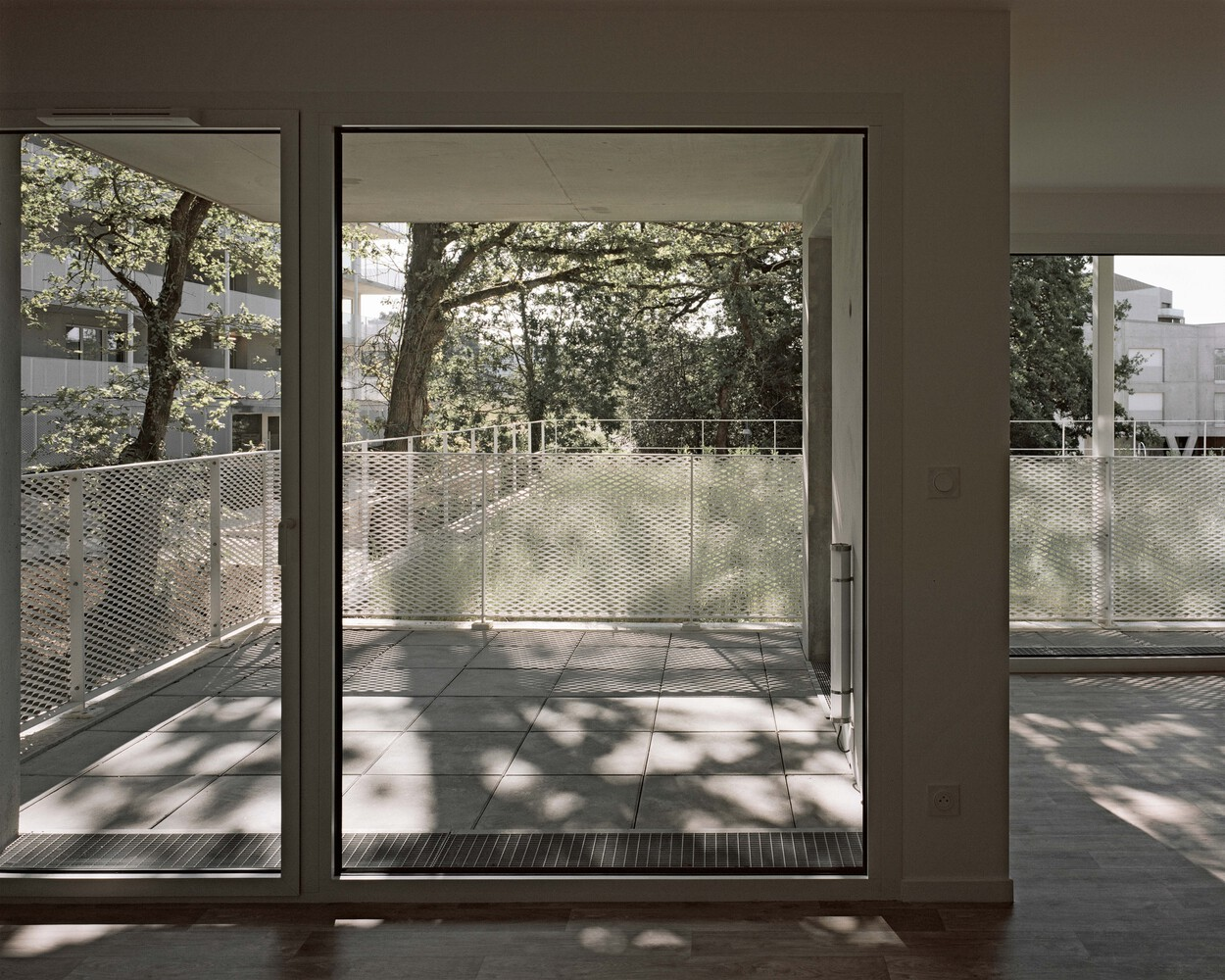 Interior view through sliding glass doors to a balcony with perforated metal railing and dappled sunlight