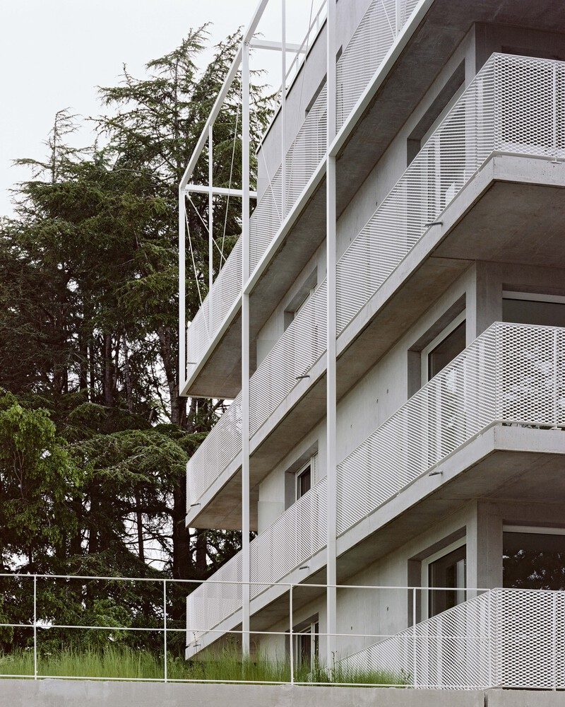Stacked concrete balconies with perforated metal screens and white diagonal bracing beside conifers