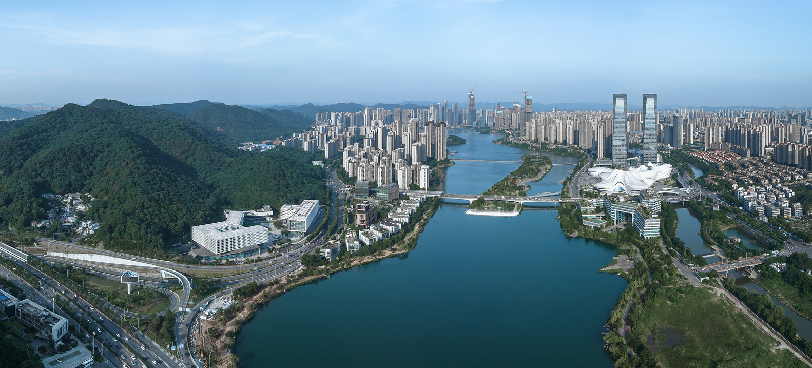 Wide aerial panorama showing the campus buildings positioned between the river and green mountain slopes