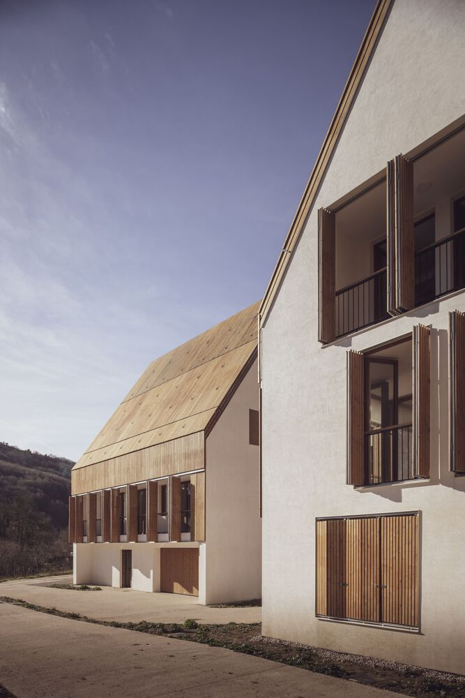 Two gabled volumes with white plaster walls and vertical timber shutters on a sloped paved street